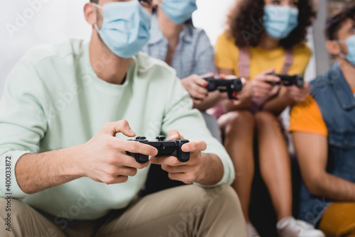Cropped view of man in medical mask holding joystick near friends on blurred background at home