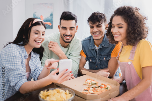Cheerful hispanic friends looking at smartphone on blurred foreground near potato chips and pizza