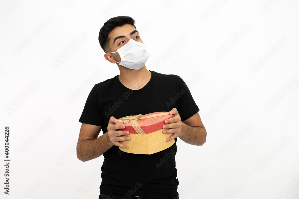 Young male model wearing a medical face mask. He is holding a gift box in the shape of a heart. He is wearing a black shirt. Isolated image and white background. Young male model wearing a medical face mask. He is holding a gift box in the shape of a heart. He is wearing a black shirt. Isolated image and white background.