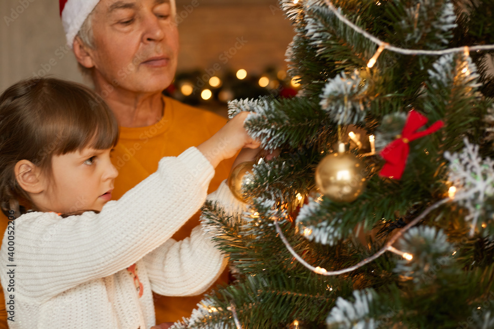 Happy family decorating xmas tree, grandfather wearing orange sweater and little girl in white jumper posing in living room, beautiful xmas tree with bubbles and bows.