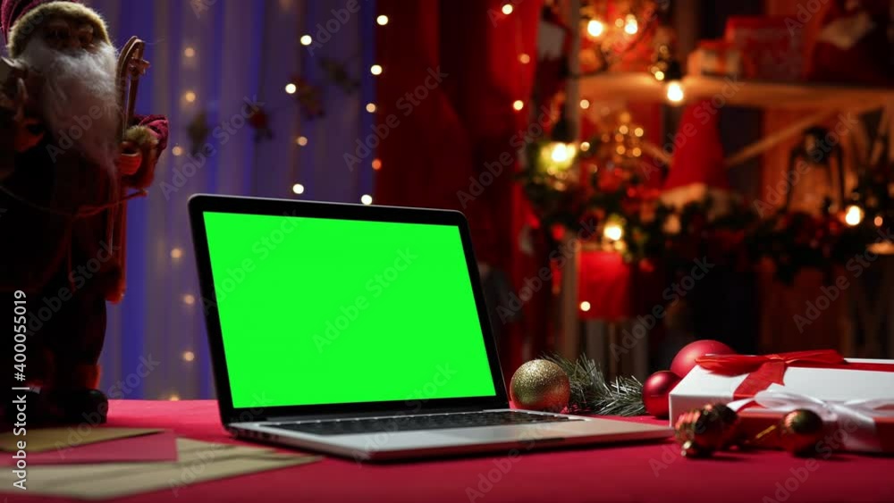 Laptop with green screen chroma key stands on red table next to Santa Claus, gifts and toys. Home room with Christmas decor and lights in the blurred background. Mockup monitor. Close up. Slow motion. Laptop with green screen chroma key stands on red table next to Santa Claus, gifts and toys. Home room with Christmas decor and lights in the blurred background. Mockup monitor. Close up. Slow motion.