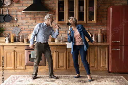 Happy older senior loving family couple having fun in kitchen  singing favorite karaoke songs in utensils. Positive joyful middle aged spouse enjoying funny domestic weekend activity together indoors.