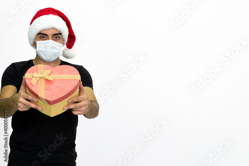 Young male wearing a medical face mask and a santa hat. Young man is holding a heart-shaped gift box in his hands. Isolated image and white background.