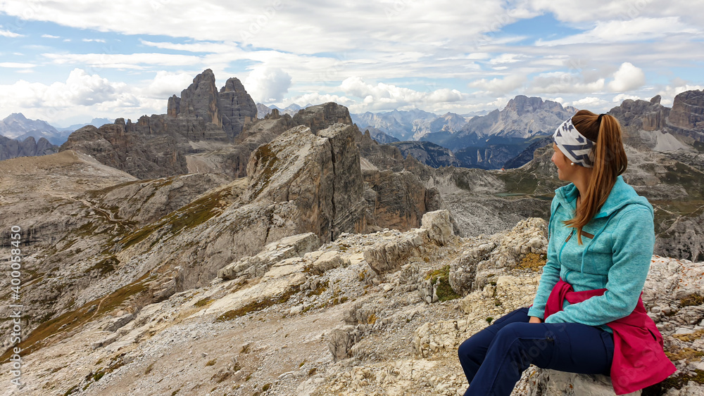 Woman hiking in high and desolated mountains in Italian Dolomites. She sits on a big boulder and enjoys the idyllic landscape. Raw and unspoiled landscape. Clear and sunny day. Endless mountain chain