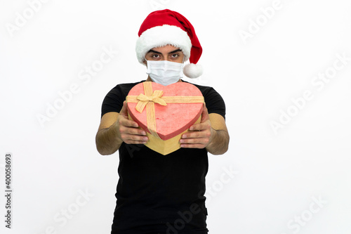 Young man wearing a Santa hat. She is holding a heart-shaped box in her hand, wearing a medical face mask.