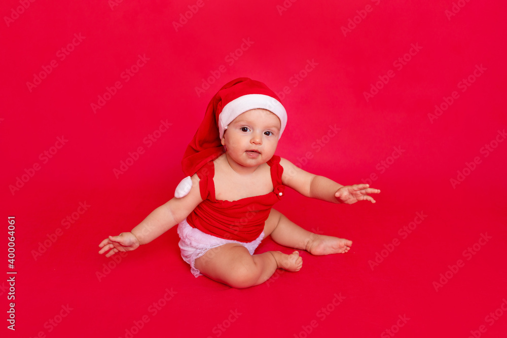 a small child of six months in a Santa hat sits on a red isolated photophone holding his hands to the sides, a place for text, the concept of the new year and holiday a small child of six months in a Santa hat sits on a red isolated photophone holding his hands to the sides, a place for text, the concept of the new year and holiday
