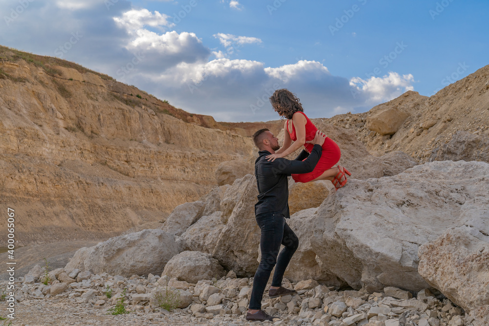 a strong young guy helps a girl in a red dress get off a big stone