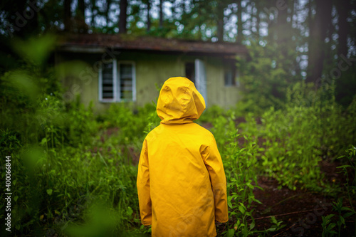 A child in a yellow coat standing in the woods in front of an old wooden house