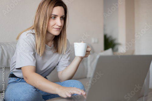 Beautiful young woman working on laptop computer while sitting at the living room, drinking coffee.