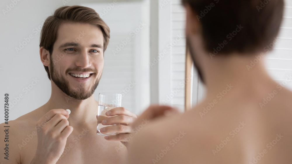 Head shot close up smiling young man looking in mirror, holding glass of water and pills, taking morning medicine or healthcare vitamins, improving immunity every day, feeling healthy and energetic. - obrazy, fototapety, plakaty 