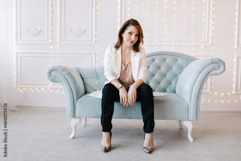 Attractive young woman in a business suit with white jacket, black pants and high heels sits on a blue classic sofa. White background with light Attractive young woman in a business suit with white jacket, black pants and high heels sits on a blue classic sofa. White background with light