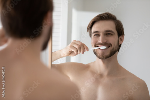 Mirror reflection happy young 30s bare european guy brushing teeth with tooth brush and toothpaste, enjoying oral dental morning cleaning routine in bathroom, involved in whitening teeth procedure.