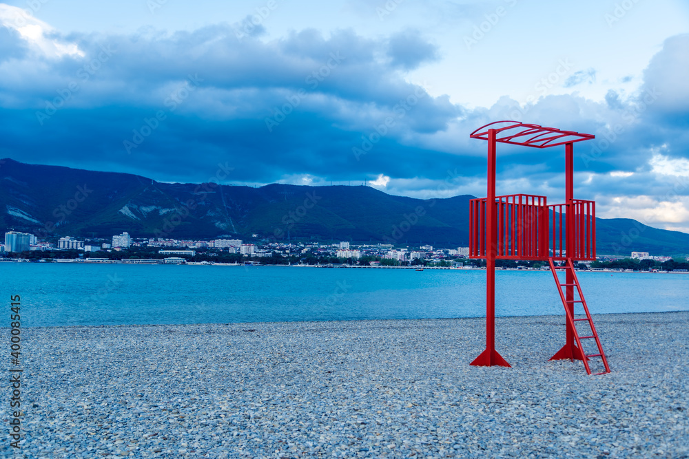 the red lifeguard tower on the pebble beach of the resort town is out of season. No people, empty beach. Winter, clouds. Mountains in the background the red lifeguard tower on the pebble beach of the resort town is out of season. No people, empty beach. Winter, clouds. Mountains in the background