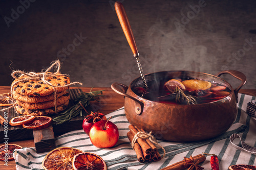 Hot mulled wine in a copper saucepan, a stack of homemade cookies, apples, slices of dry orange, cinnamon sticks, a tablecloth against the background of an old wooden background.