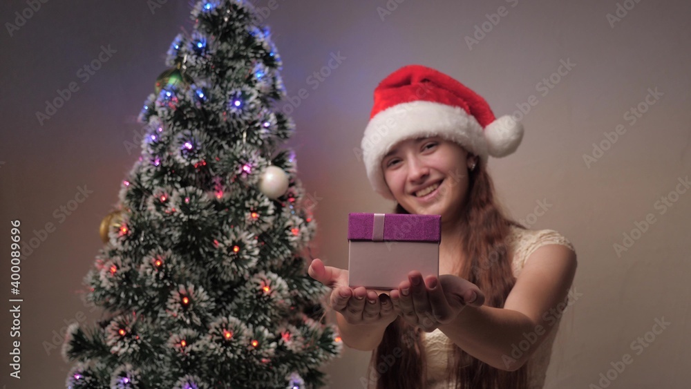 A girl in a Santa Claus hat is holding gift next to Christmas tree. Teenager holds out gift box to camera, rejoices and smiles. Family children's holiday and celebration, winter rest. Merry Christmas.