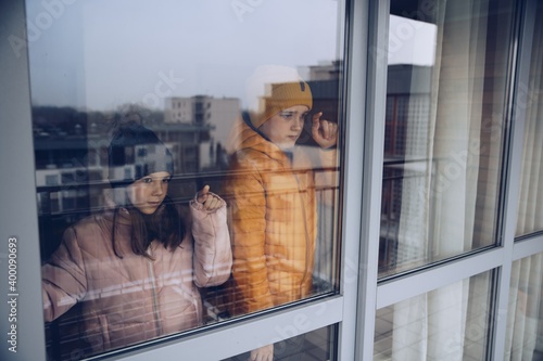 Two children are standing by window and sadly looking outside during quarantine or isolation due to pandemic. photo