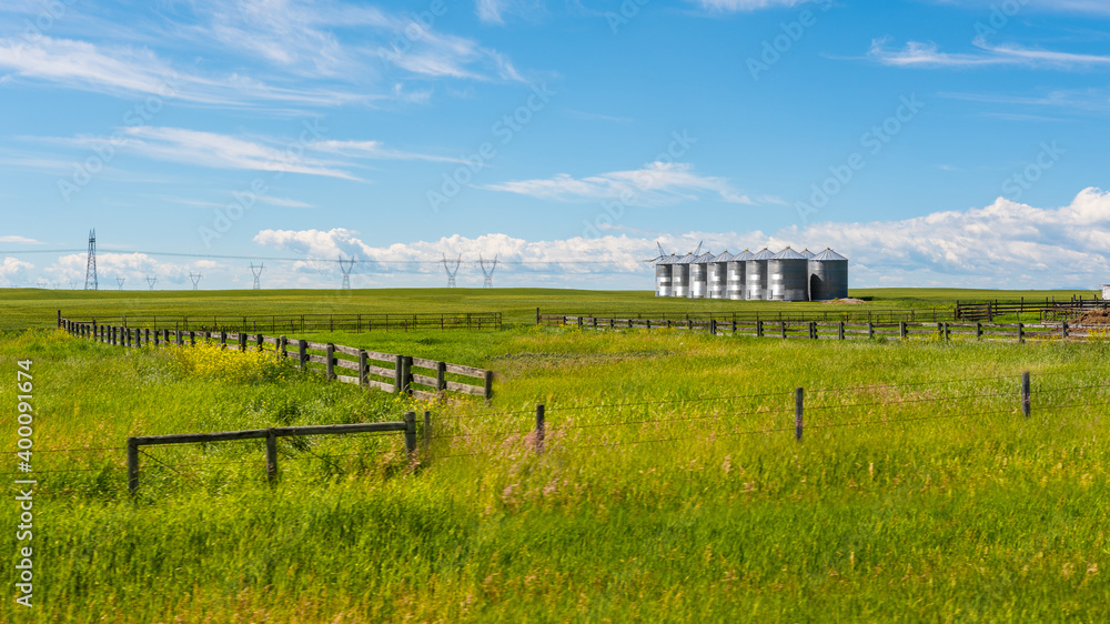 Grain silos in grassy field farmland and wooden fence Alberta prairie landscape background. Agriculture and farming industry. Rural prairies grassland and beautiful blue sky