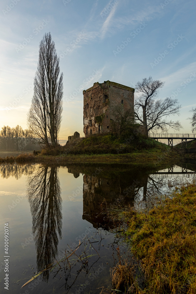Winter barren trees and the ruins of Nijenbeek castle reflecting in the spill water of river IJssel in The Netherlands, backlit by an early morning sunrise
