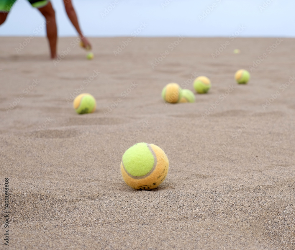 Many beach game balls on the beach, selective perspective with partial view of a person reaching for one ball in blurred background.
