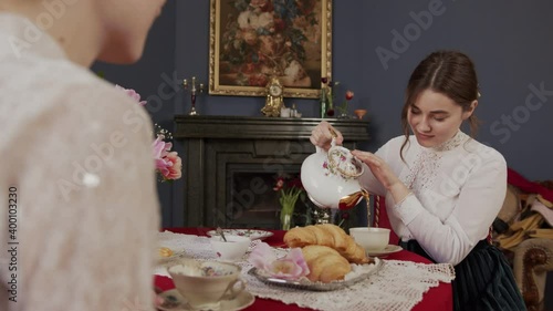 very beautiful girl in a vintage white shirt sits at a beautiful antique table and pours herself tea