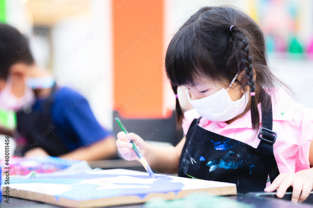 Child wearing cloth mask is in an art class. Girl are working on painting watercolor on the canvas. Children grabs the paintbrush with his right hand. New normal. Prevent spreading the coronavirus. Child wearing cloth mask is in an art class. Girl are working on painting watercolor on the canvas. Children grabs the paintbrush with his right hand. New normal. Prevent spreading the coronavirus.