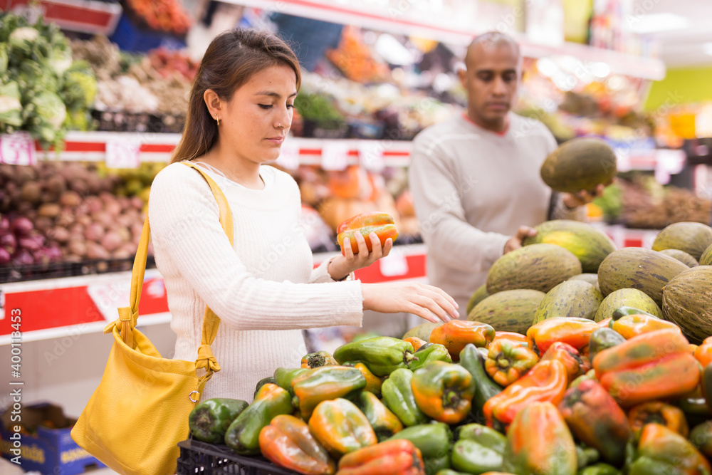 Young latin american girl choosing sweet pepper in supermarket with her boyfriend