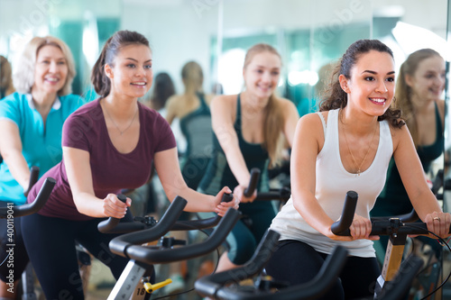 Cheerful females of different age cycling on exercise bikes at fitness club .