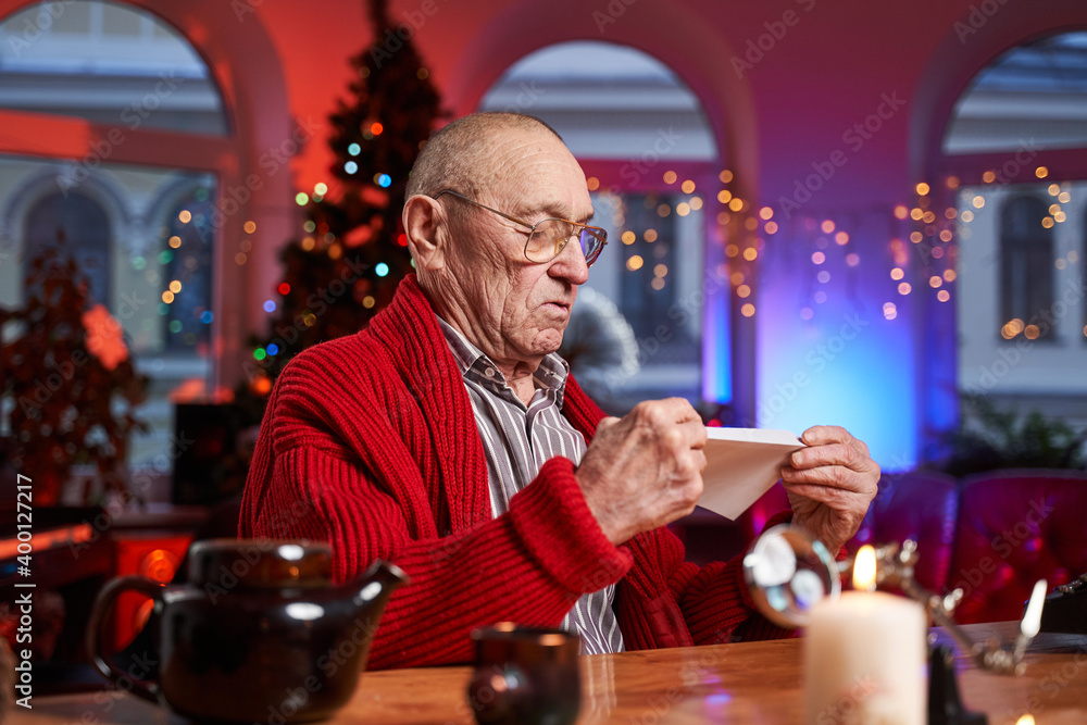 Bald and serious elder man sitting at table and holding a letter in cosy and decorated room with christmas tree.