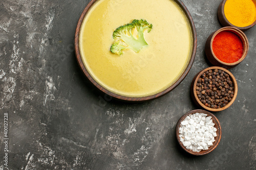 Horizontal view of creamy broccoli soup in a brown bowl and different spices on gray table