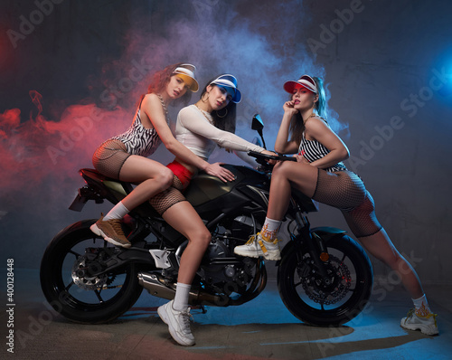 Beautiful slim girls in short clothing with caps posing on motorbike in smokey and colourful garage.