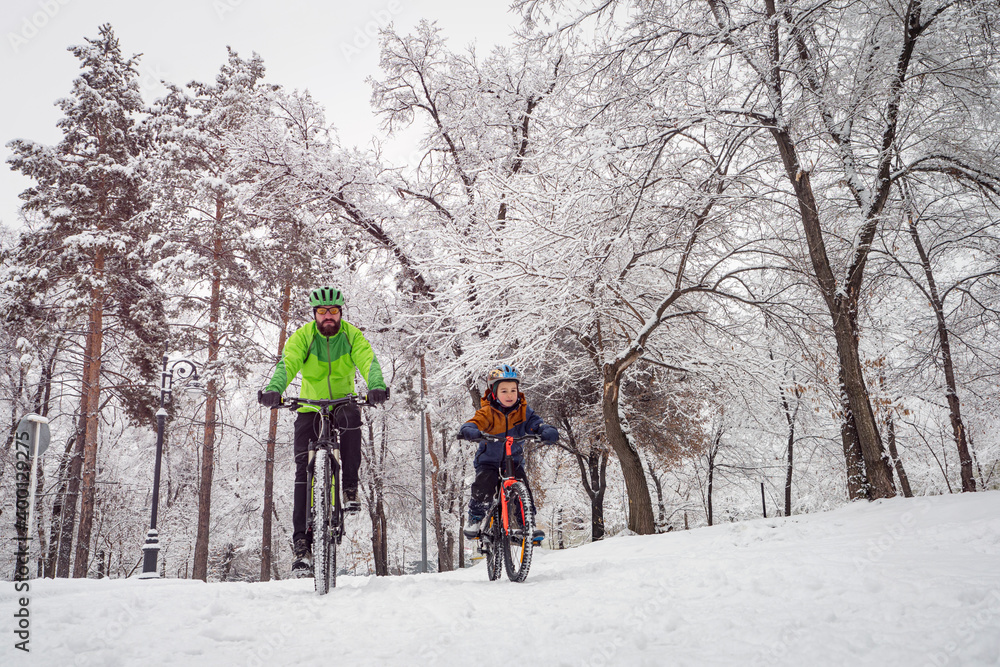 Father and son ride bicycles in the winter park. Bearded man and boy on bicycles spend family weekend in beautiful snowy winter park