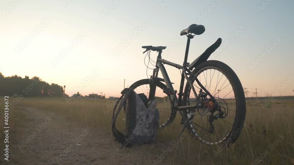 Bicycle Stands in the Field, in the Background Forest and Sky. There is a Backpack near the Bicycle. Summer Landscape with Cycling Concept. Bicycle Stands in the Field, in the Background Forest and Sky. There is a Backpack near the Bicycle. Summer Landscape with Cycling Concept.