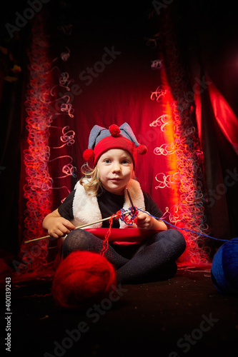 A pretty little girl who looks like a dwarf or elf is knitting with multicolored threads from large clubs on a black and red background.