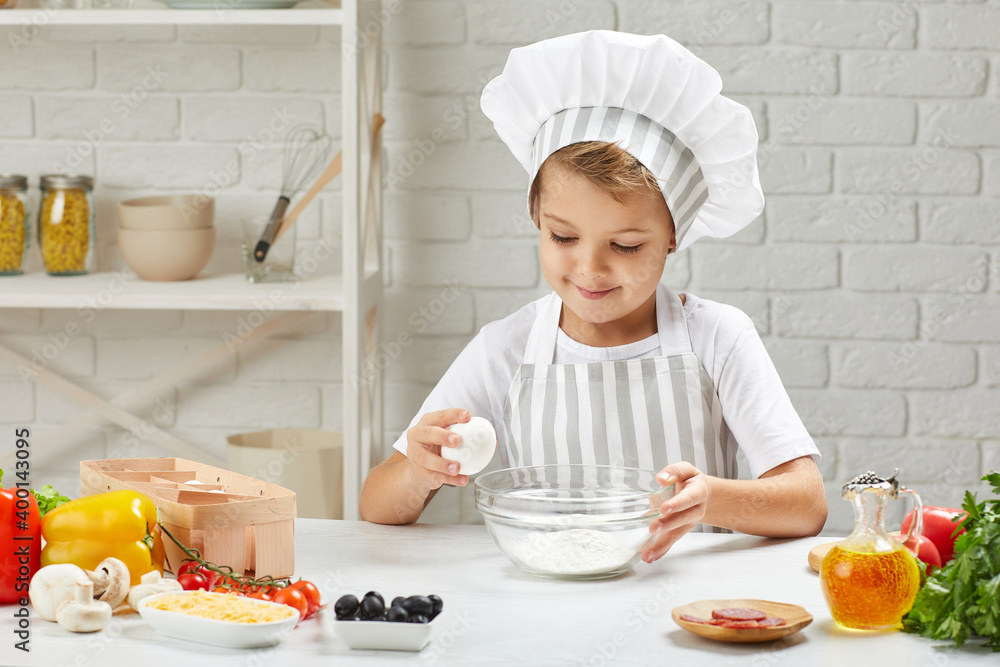 little boy in chef hat and an apron is breaking eggs for making pizza in the kitchen. child cooking with dough little boy in chef hat and an apron is breaking eggs for making pizza in the kitchen. child cooking with dough