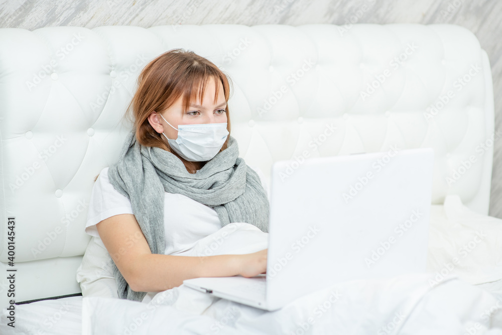 Teen girl wearing medical protective mask sits on a bed at home and uses laptop for education. Quarantine and coronavirus epidemic concept