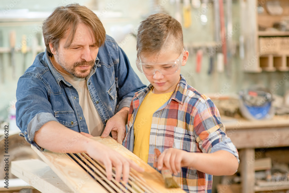 Father teaches his young son how to paint a wooden board with a paintbrush in a carpentry workshop Father teaches his young son how to paint a wooden board with a paintbrush in a carpentry workshop