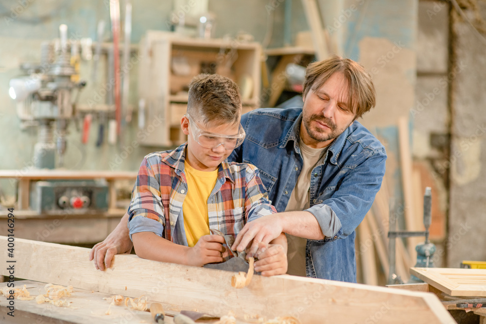 Father teaches his young son to plan wood in a carpentry workshop Father teaches his young son to plan wood in a carpentry workshop