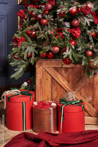 red boxes with gifts, round in shape, tied with a green ribbon, stand under a Christmas decorated tree