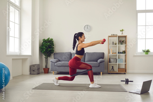 Side view of a woman training with dumbbells and rubber bands at home and using a laptop.