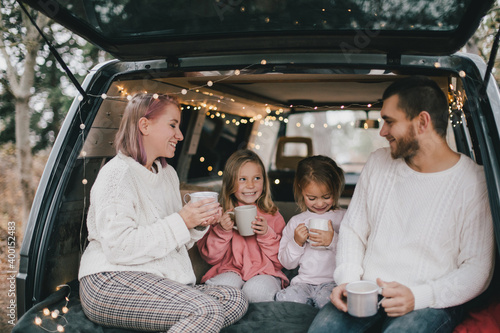 Happy parents and their kids drinking hot cocoa sitting in a van decorated with festive Christmas lights. © polinaloves