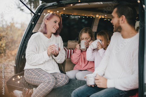 Happy parents and their kids drinking hot cocoa sitting in a van decorated with festive Christmas lights.