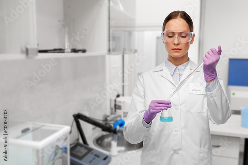 science, chemistry and people concept - young female scientist in goggles with pipette chemical in flask making test or research over laboratory background