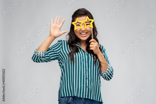party props, photo booth and people concept - happy asian young woman with big glasses in shape of stars waving hand over grey background