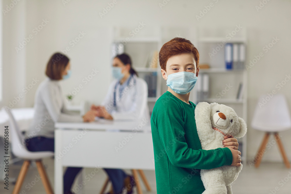 Portrait of happy boy in face mask hugging his toy, standing against blurred hospital office