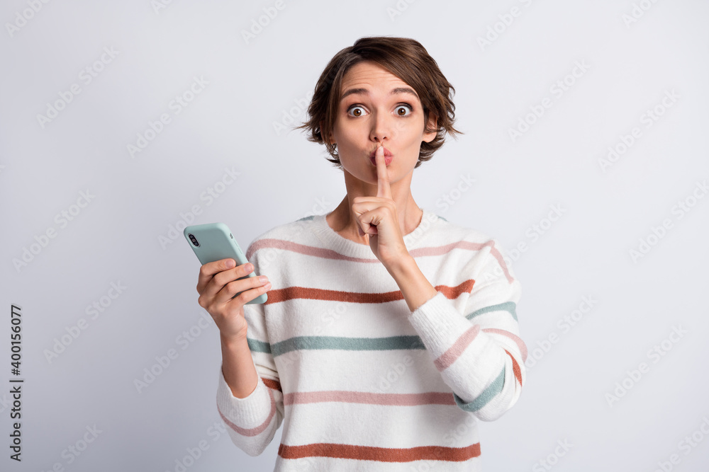Photo of funny young lady wear striped sweater holding modern gadget ask not tell secrets isolated grey color background Photo of funny young lady wear striped sweater holding modern gadget ask not tell secrets isolated grey color background