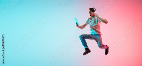 Music, drive. Young caucasian man's jumping on gradient blue-pink studio background in neon light. Concept of youth, human emotions, facial expression, sales, ad. Half length, copyspace. Flyer