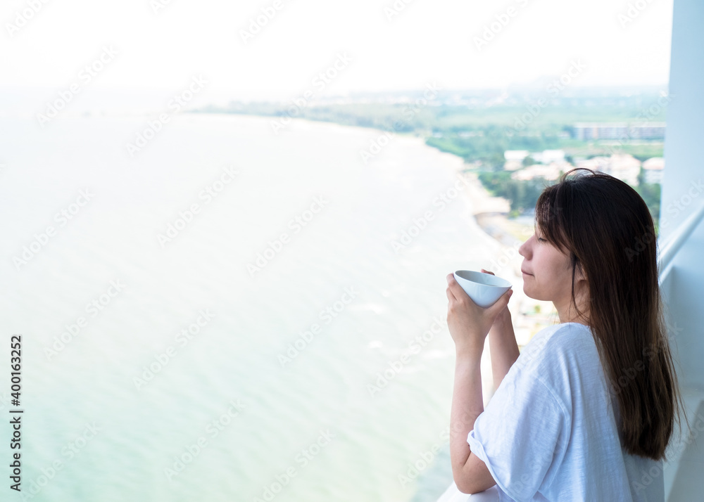Asian Woman drinking hot drink in morning at balcony for breakfast. Beautiful cozy warm lady having white coffee cup on luxury hotel terrace with sea view in summer travel holidays.