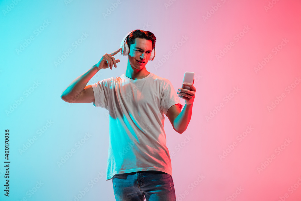 Listening to music, dancing. Young caucasian man's portrait on gradient blue-pink studio background in neon. Concept of youth, human emotions, facial expression, sales, ad. Half length, copyspace.