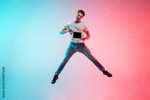 Tablet screen. Young caucasian man's jumping on gradient blue-pink studio background in neon light. Concept of youth, human emotions, facial expression, sales, ad. Full length, copyspace.