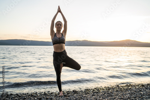 Young healthy girl standing in yoga tree pose by the lake